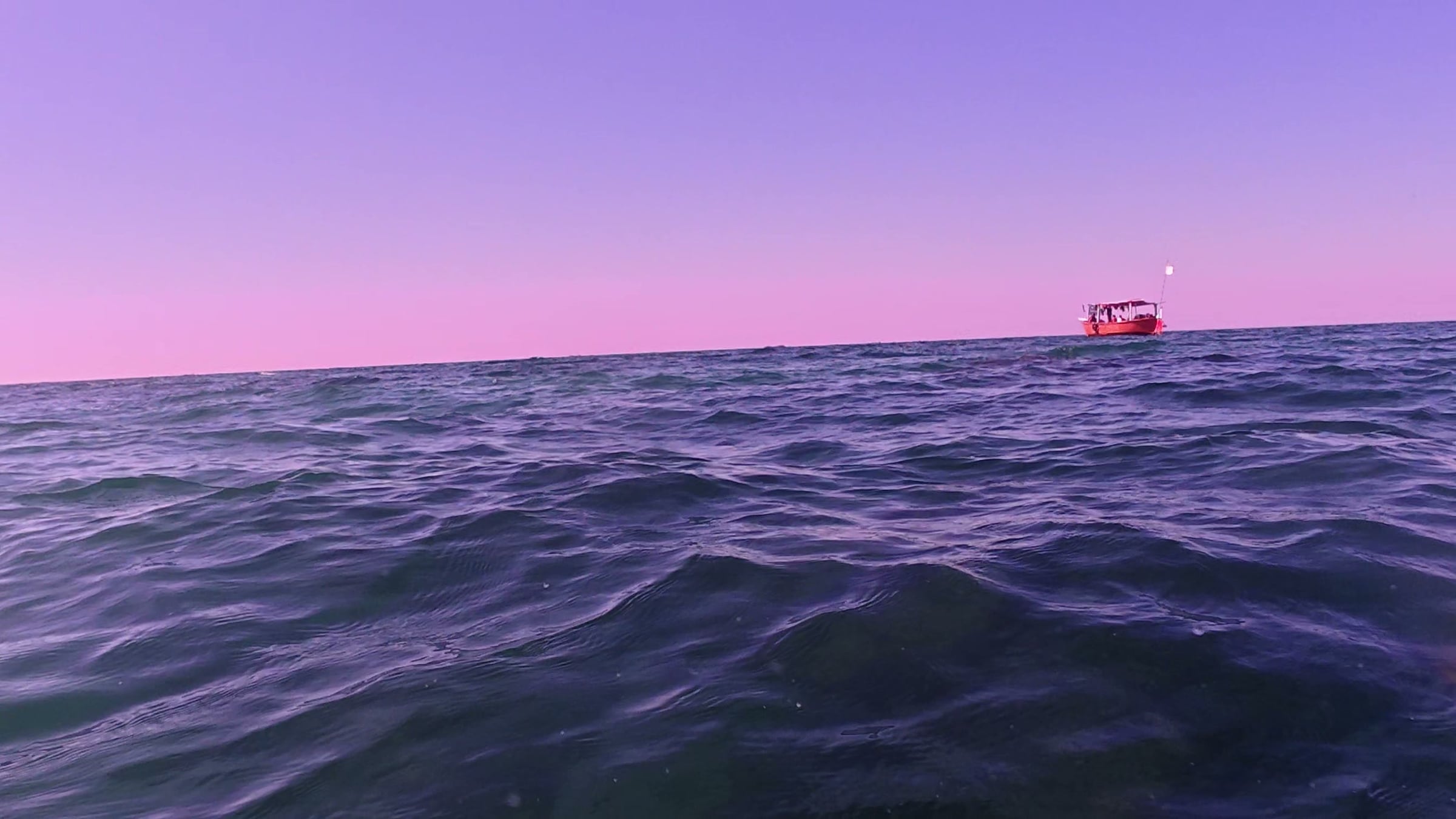 A small red fishing boat alone on the horizon at first light, photographed at water level, the Arabian Sea violet under a pink sky